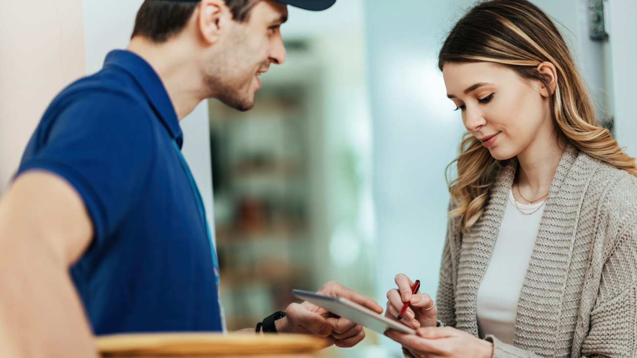 Young woman using touchpad to sign for package delivery from a courier while standing on a doorway.